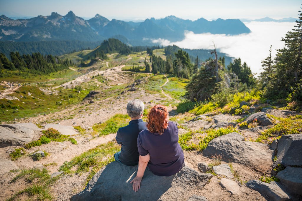 Two senior adults sitting on rocks, admiring a stunning mountain landscape.